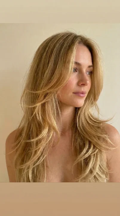 A woman with long feathered layers showing soft wispy ends fanning outward with natural movement and dimension, one of the most timeless yet freshly relevant haircut trends for 2026, photographed in golden blonde tones under warm afternoon window light.